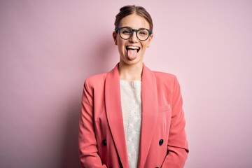 Young beautiful redhead woman wearing jacket and glasses over isolated pink background sticking tongue out happy with funny expression. Emotion concept.