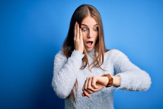 Young beautiful redhead woman wearing casual sweater over isolated blue background Looking at the watch time worried, afraid of getting late