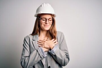 Young beautiful redhead architect woman wearing security helmet over white background smiling with hands on chest with closed eyes and grateful gesture on face. Health concept.