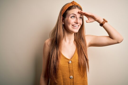 Young beautiful redhead woman wearing casual t-shirt and diadem over yellow background very happy and smiling looking far away with hand over head. Searching concept.
