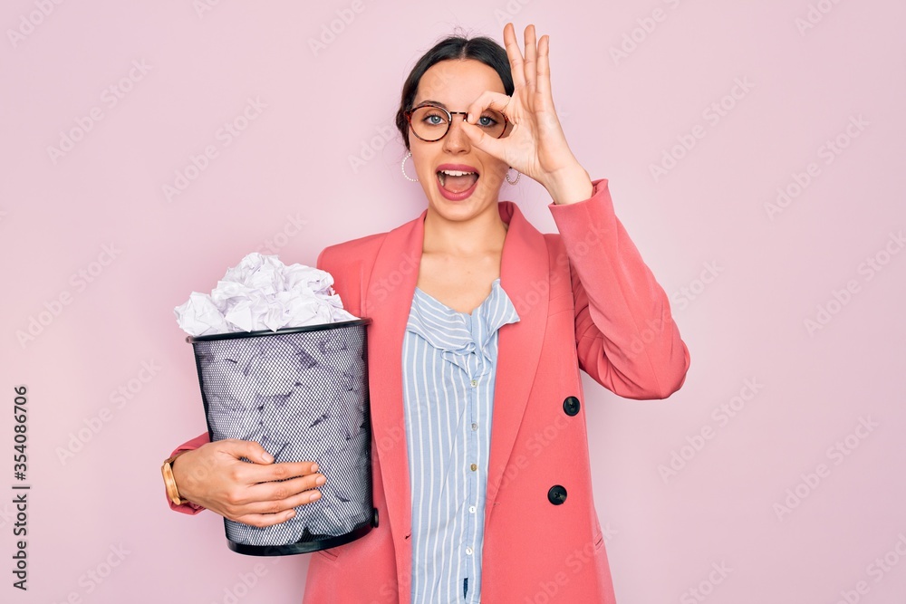 Wall mural Young beautiful business woman with blue eyes holding heap bin of crumpled papers with happy face smiling doing ok sign with hand on eye looking through fingers