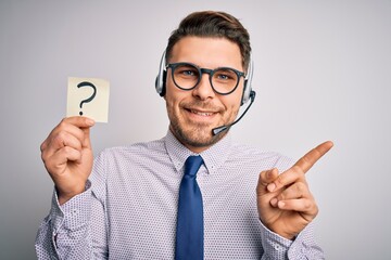 Young call center operator business man with blue eyes holding paper note with question mark very happy pointing with hand and finger to the side