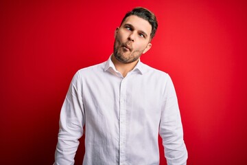 Young business man with blue eyes wearing elegant shirt standing over red isolated background making fish face with lips, crazy and comical gesture. Funny expression.