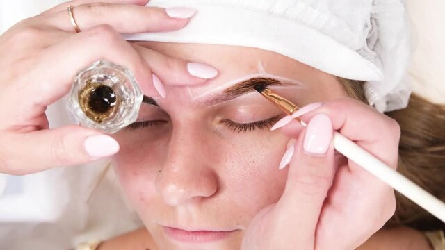 Permanent make-up for eyebrows of woman with thick brows in beauty salon. Closeup beautician doing coloring tattooing eyebrow.