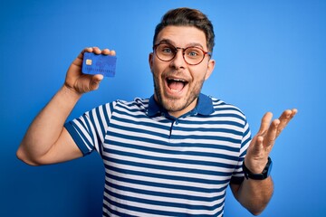 Young man with blue eyes wearing glasses and holding credit card over blue background very happy and excited, winner expression celebrating victory screaming with big smile and raised hands