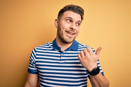 Young man with blue eyes wearing casual striped t-shirt over yellow background smiling with happy face looking and pointing to the side with thumb up.