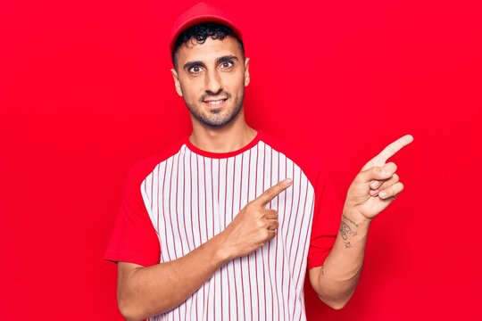 Young hispanic man wearing baseball uniform smiling and looking at the camera pointing with two hands and fingers to the side.