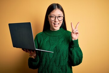 Young asian business woman wearing glasses and working using computer laptop smiling with happy face winking at the camera doing victory sign with fingers. Number two.