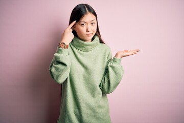 Young beautiful asian woman wearing green winter sweater over pink solated background confused and annoyed with open palm showing copy space and pointing finger to forehead. Think about it.