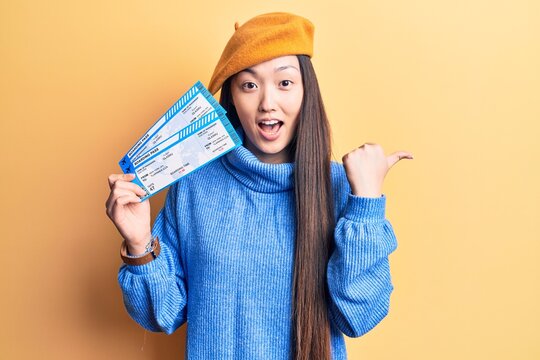 Young beautiful blonde woman holding airline boarding pass pointing thumb up to the side smiling happy with open mouth
