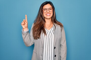 Young hispanic business woman wearing glasses standing over blue isolated background showing and pointing up with finger number one while smiling confident and happy.