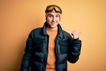 Young handsome caucasian man wearing hat, coat and ski glasses for winter and snow weather smiling with happy face looking and pointing to the side with thumb up.