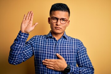 Young handsome latin man wearing casual shirt and glasses over yellow background Swearing with hand on chest and open palm, making a loyalty promise oath