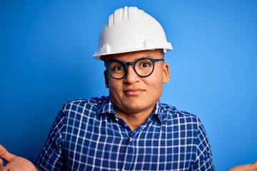 Young handsome engineer latin man wearing safety helmet over isolated blue background clueless and confused expression with arms and hands raised. Doubt concept.