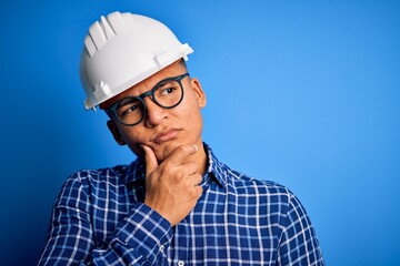 Young handsome engineer latin man wearing safety helmet over isolated blue background with hand on chin thinking about question, pensive expression. Smiling with thoughtful face. Doubt concept.