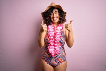 Young beautiful arab woman on vacation wearing swimsuit and hawaiian lei flowers success sign doing positive gesture with hand, thumbs up smiling and happy. Cheerful expression and winner gesture.