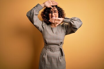 Beautiful arab business woman wearing dress and glasses standing over yellow background Smiling cheerful playing peek a boo with hands showing face. Surprised and exited