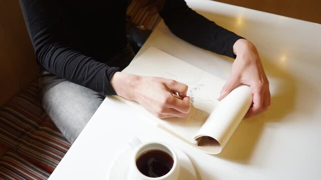 a Hand of a girl draws in sketchbook with a pencil in a cafe, a cup of coffee on the table, Camera shifting in from the top in spiral