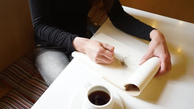a Hand of a girl draws in sketchbook with a pencil in a cafe, a cup of coffee on the table, Camera shifting in from the top
