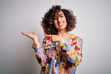 Young beautiful curly arab woman wearing floral colorful shirt standing over white background amazed and smiling to the camera while presenting with hand and pointing with finger.