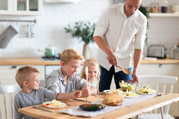 Obraz na płótnie Canvas family father with three kids two sons and daughter eating healthy food in kitchen at home, dad puts green salad on plates to his kids.