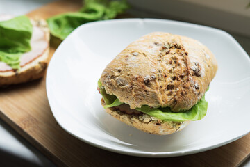 Sandwich with lettuce and ham on white plate. Wooden kitchen board on white table. Lettuce in the background.