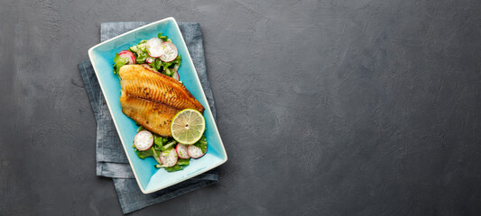 Grilled fish sea bass fillet with green lettuce and radish. Black stone background. Top view with...