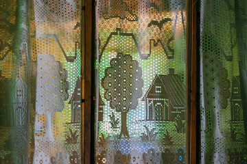 Vintage white openwork transparent curtains with houses, trees, plants, flowers, birds, clouds pattern in front of the light on the old wooden window frame. Background - green, orange, blue. Closeup