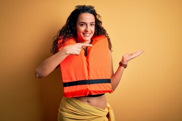 Young beautiful woman with curly hair wearing orange lifejacket over yellow background amazed and smiling to the camera while presenting with hand and pointing with finger.
