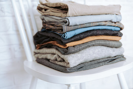 Stack of neatly folded casual clothes on white chair. Pile of ironed beige and grey color cotton clothes and sweaters. Order and minimalism concept