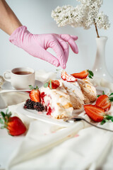 A gloved hand decorates a meringue cake, garnished with nut chips and strawberries, among lilac flowers. Food photography. Advertising and commercial close up design.
