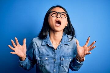 Young beautiful chinese woman wearing casual denim shirt over isolated blue background crazy and mad shouting and yelling with aggressive expression and arms raised. Frustration concept.