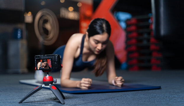 The camera on the tripod is taking pictures or videos. Asian Women Trainer In Good Shape Teaching or performing a sample of plank poses is a bodyweight exercise in online training concept.