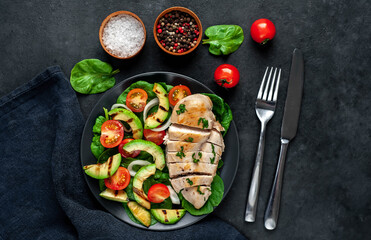 
Grilled chicken breast and grilled avocado salad with cherry tomatoes, spinach, in a black plate on a stone background