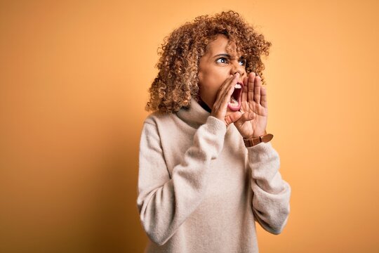 Young beautiful african american woman wearing turtleneck sweater over yellow background Shouting angry out loud with hands over mouth
