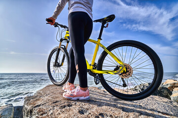 Fototapeta na wymiar Fitness woman with bicycle stands on the stones on the seashore against the blue sky. Athletic healthy people with active and sporty lifestyle