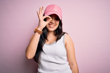 Naklejka premium Young brunette woman wearing casual sport cap over pink background doing ok gesture with hand smiling, eye looking through fingers with happy face.