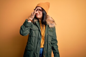 Young brunette woman wearing glasses and winter coat with hat over yellow isolated background smelling something stinky and disgusting, intolerable smell, holding breath with fingers on nose