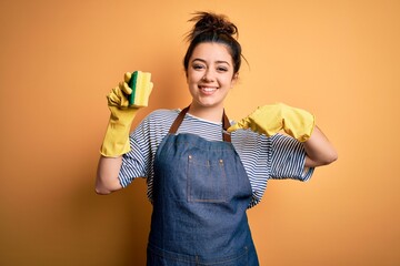 Young brunette cleaner woman wearing housekeeping gloves holding scourer scrub with surprise face...
