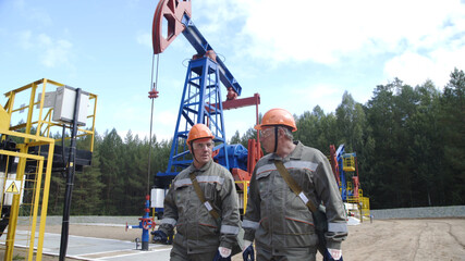 Two man oil workers in orange helmets walking and talking near oil pump jacks. Oil engineers overseeing site of crude oil production. Balancing drive rod pumps oil rocker.
