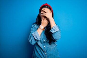 Young brunette woman wearing casual denim shirt over blue isolated background Covering eyes and mouth with hands, surprised and shocked. Hiding emotion