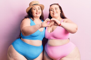 Young plus size twins wearing bikini smiling in love showing heart symbol and shape with hands....