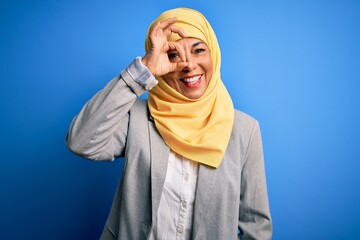 Middle age brunette business woman wearing muslim traditional hijab over blue background doing ok gesture with hand smiling, eye looking through fingers with happy face.