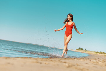 Fototapeta na wymiar Young attractive brunette woman in red body running on a coast with blue sky on background