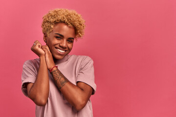 Happy cheerful african american woman rejoicing gift, keeps hands together near face, focused away, notices desirable things, laughs on a pink background. Personal offers and discounts in online store