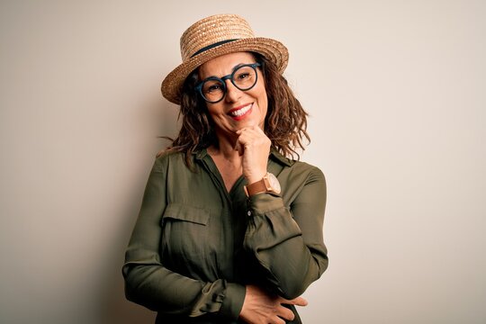 Middle age brunette woman wearing glasses and hat standing over isolated white background looking confident at the camera with smile with crossed arms and hand raised on chin. Thinking positive.