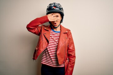 Middle age motorcyclist woman wearing motorcycle helmet and jacket over white background peeking in shock covering face and eyes with hand, looking through fingers with embarrassed expression.