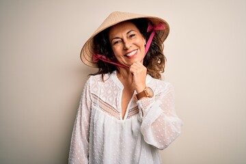 Middle age brunette woman wearing asian traditional conical hat over white background looking confident at the camera with smile with crossed arms and hand raised on chin. Thinking positive.