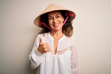 Middle age brunette woman wearing asian traditional conical hat over white background doing happy thumbs up gesture with hand. Approving expression looking at the camera showing success.