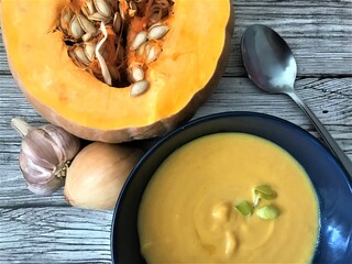 Dark blue bowl with pumpkin puree soup  with a spoon on the right, untreated head of garlic and onion on the left and cut pumpkin with seeds on top against the background of old gray wood
